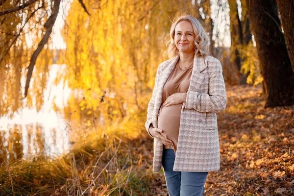 Portrait of middle-aged pregnant woman outdoors at park. Middle age pregnant woman expecting baby at aged pregnancy. Blonde woman wearing brown sweater and beige coat.