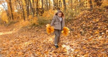 Little kid in a stylish costume having fun on the weekend. The girl is holding bunch of leaves and running in the beautiful autumn park.