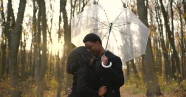 Lovely black couple cuddling while date in autumn park. The guy holding umbrella.
