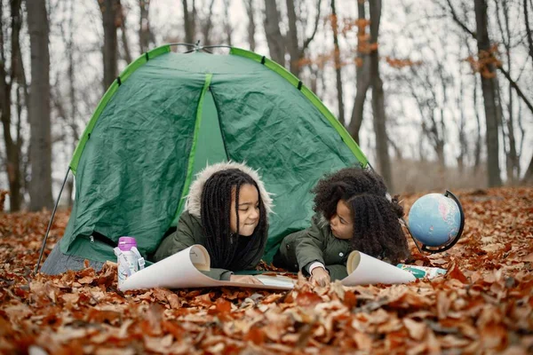 Beautiful little black girls in tent camping in the forest. Two little sisters lying in a tent in autumn forest and looking on a map. Black girls wearing khaki coats.