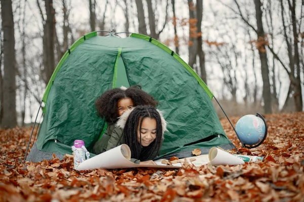Beautiful little black girls in tent camping in the forest. Two little sisters lying in a tent in autumn forest and looking on a map. Black girls wearing khaki coats.