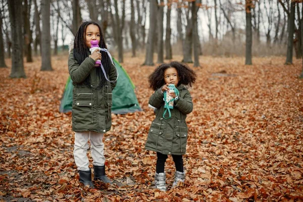 美丽的黑人小女孩站在森林的帐篷附近 两个小妹妹在秋天的森林里喝茶 穿着卡其布外套的黑人女孩 — 图库照片
