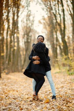 Siyah genç adam ve kız arkadaşı sarılıyor. Sonbahar parkında yürüyen romantik bir çift. Siyah paltolu kadın ve erkek..
