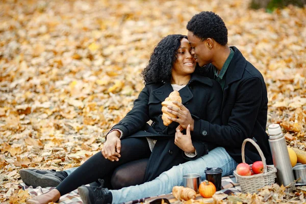 Black man and woman hugging and eating a croissant. Romantic couple sitting on a blanket in autumn park. Man and woman wearing black coats.