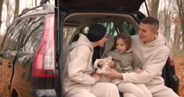 Mutlu genç bir aile, sonbahar ormanında dışarıda geçirilen bir günün ardından dinleniyor. Ebeveynler küçük kızlarıyla kucaklaşıyorlar ve arabalarının bagajında Fransız buldogları var..