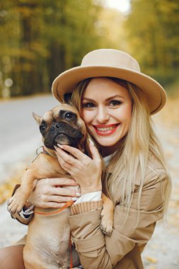 Kadınlar ve köpekler sonbahar parkında oynayıp eğleniyorlar. Kahverengi buldog ve bayan sahibi parkta bir gün yürüyorlar. Bej ceketli ve şapkalı bir kadın..