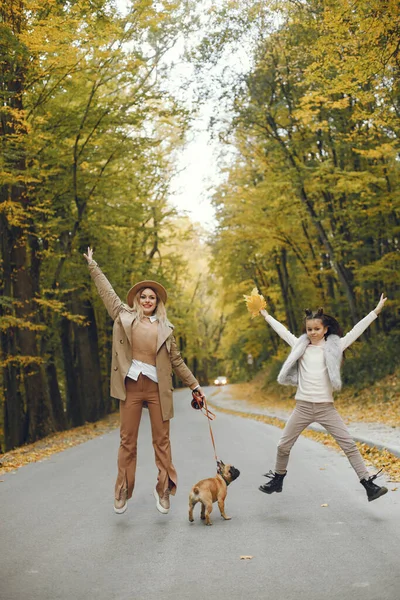 Woman, little girl and dog walking in the autumn park. Brown french bulldog with female owners spend a day at the park playing and having fun. Woman wearing beige coat and a hat.