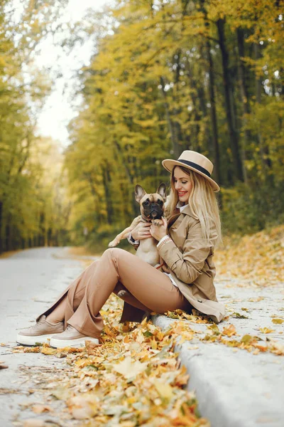Woman and dog play and have fun in the autumn park. Brown french bulldog with female owner spend a day at the park playing and having fun. Woman wearing beige coat and a hat.