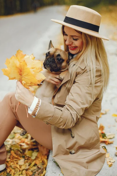 Woman and dog play and have fun in the autumn park. Brown french bulldog with female owner spend a day at the park playing and having fun. Woman wearing beige coat and a hat.