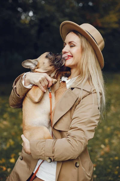 Woman and dog play and have fun in the autumn park. Brown french bulldog with female owner spend a day at the park playing and having fun. Woman wearing beige coat and a hat.