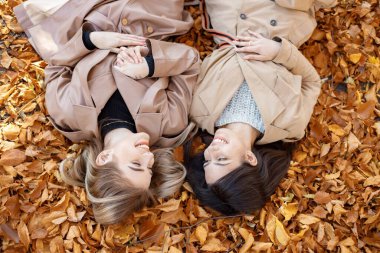 Sonbahar parkında sarı yaprakların üzerinde yatan iki kız. Bayan arkadaşlarının portresi. Bej ceketli iki kız..