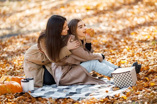 两个漂亮的女性朋友花时间在草地上的野餐毛毯上 两个笑着的妹妹在秋天的公园里野餐吃羊角面包 穿着外套的布鲁内特和金发女孩 — 图库照片