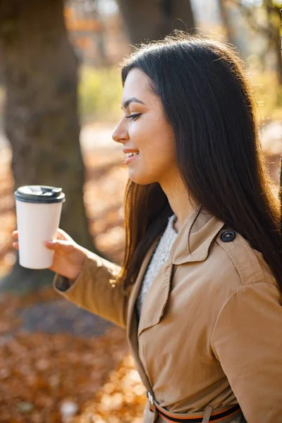 Beautiful woman drink coffee and posing for the camera in autumn park. Young girl standing near tree with a coffee. Brunette woman wearing beige coat.