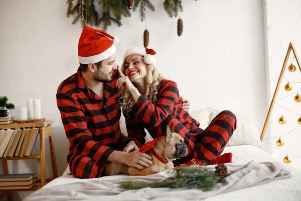 Lovely couple celebrating christmas with dog on a bed. Blonde woman and brunette man wearing plaid pajamas. French bulldog with his family.