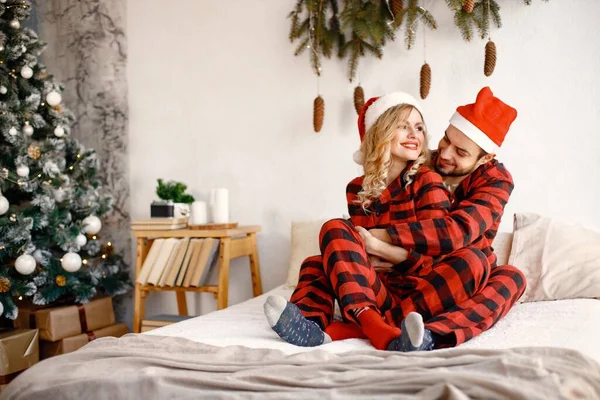 Lovely couple celebrating christmas on a bed. Blonde woman and brunette man wearing plaid pajamas. Romantic couple hugging.