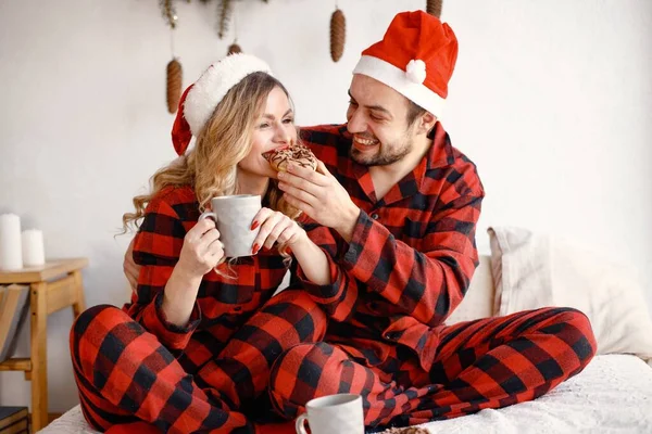 Lovely couple celebrating christmas on a bed. Blonde woman and brunette man wearing plaid pajamas. Romantic couple drinking a tea from a mug and eating a donuts.