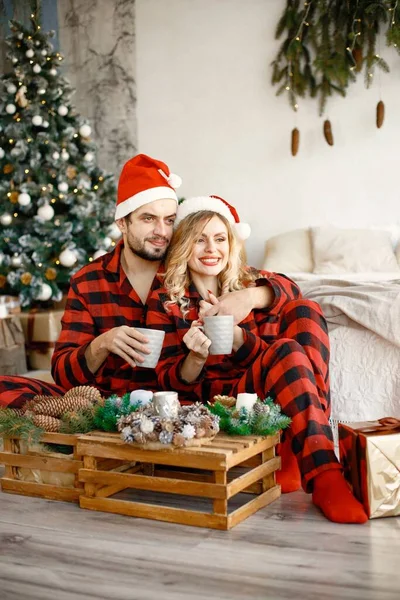 Romantic couple celebrating Christmas on a floor near Christmas tree. Blonde woman and brunette man wearing plaid pajamas. Romantic couple drinking a tea from a mugs.