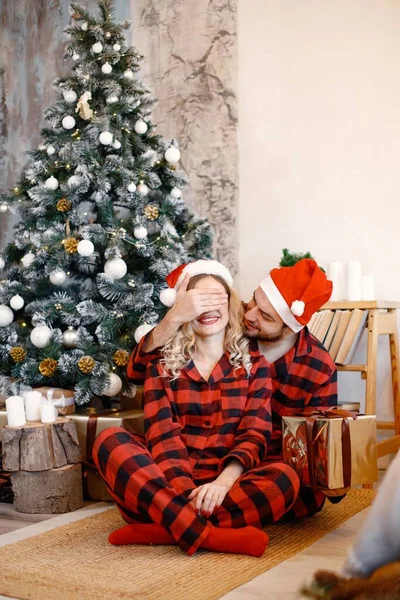 Romantic couple celebrating Christmas on a floor near Christmas tree. Blonde woman and brunette man wearing plaid pajamas. Wife and husband opening present box.