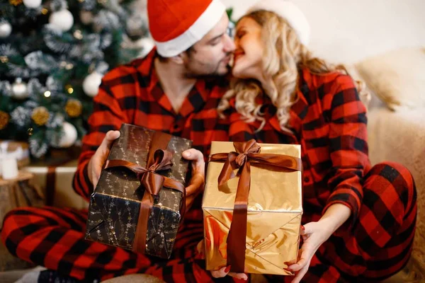 Focus on a present boxes. Romantic couple celebrating Christmas on a floor near Christmas tree. Blonde woman and brunette man wearing plaid pajamas.