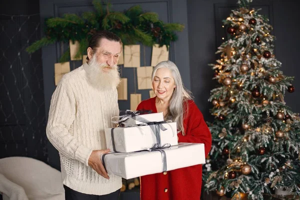 Senior man celebrating christmas with his wife near beautiful christmas tree. Old bearded man standing near his wife with grey hair and holding a gifts. Man wearing white sweater and woman red one.