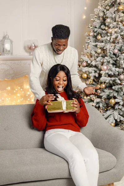 クリスマスの前夜に贈り物を持つ若い黒のカップル 女は家でコーチに座っている 男は後ろに立って彼女のボーイフレンドのための贈り物を提示しました — ストック写真