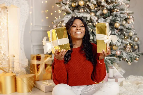 クリスマスツリーの近くに家に座っている黒人女性 女性は黄金の紙で贈り物を開ける ブルネットの女の子は赤いセーターと白いズボンを着て — ストック写真