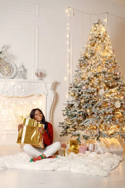 クリスマスツリーの近くに家に座っている黒人女性 女性は黄金の紙で贈り物を開ける ブルネットの女の子は赤いセーターと白いズボンを着て — ストック写真