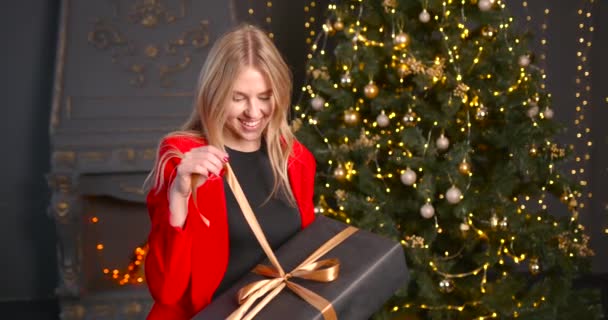 女性は手にリボン付きのクリスマスプレゼント箱を持っています 赤いジャケットを着た女性は新年を祝い モミの木の下で見つかったプレゼントを開きます 休日のコンセプト — ストック動画