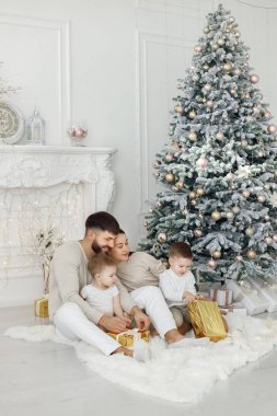 Anne, baba ve iki oğlu Noel ağacının yanında oturuyor. Heyecanlı aile Noel hediyelerini evde açıyor. Beyaz elbiseli kadın, erkek ve iki çocuk..