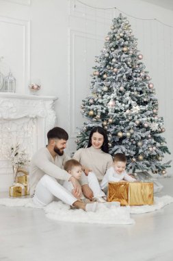 Anne, baba ve iki oğlu Noel ağacının yanında oturuyor. Heyecanlı aile Noel hediyelerini evde açıyor. Beyaz elbiseli kadın, erkek ve iki çocuk..