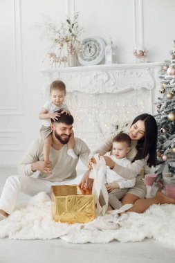 Anne, baba ve iki oğlu Noel ağacının yanında oturuyor. Heyecanlı aile Noel hediyelerini evde açıyor. Beyaz elbiseli kadın, erkek ve iki çocuk..