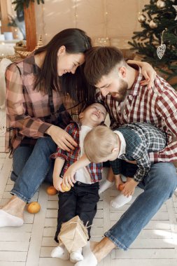 Anne, baba ve iki oğlu Noel ağacının yanında oturuyor. Ailesi olan çocuklar birlikte vakit geçiriyorlar. Kadın, erkek ve iki çocuk ekose gömlek giyiyor..