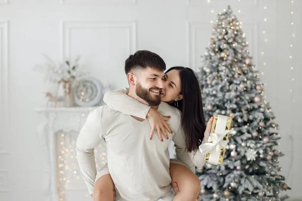 クリスマスツリーの近くでクリスマスを祝うロマンチックなカップル 白い服を着たブルネットの女性と男 女のピギーバックを持つ男 — ストック写真