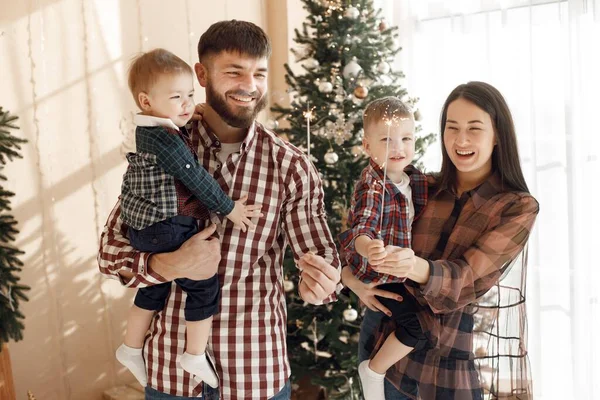 Mother, father and two their sons standing near Christmas tree. Excited family holding a sparkles. Woman, man and two boys wearing plaid shirts.