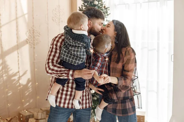 Mother, father and two their sons standing near Christmas tree. Excited family holding a sparkles. Woman, man and two boys wearing plaid shirts.