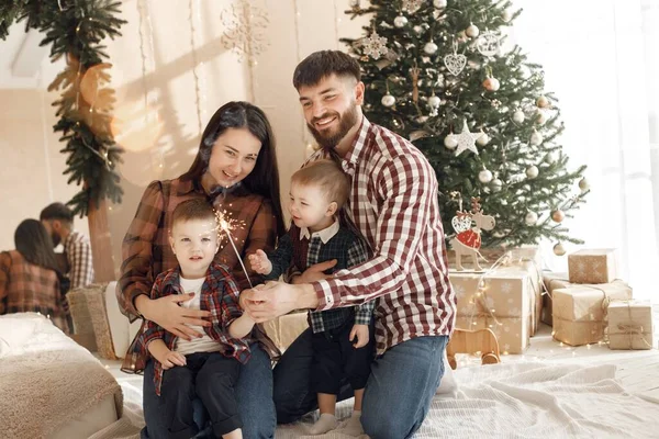 Mother, father and two their sons sitting near Christmas tree. Excited family holding a sparkle. Woman, man and two boys wearing plaid shirts.