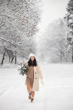 Esmer kadın ormanda durup poz veriyor. Tencerede küçük bir Noel ağacı tutan bir kadın. Kışlık kıyafetler giyen ve eldivenli şapka takan bir kız..