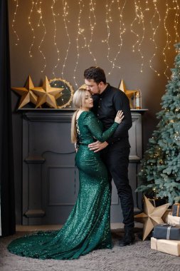 Genç bir kadın ve erkek Noel ağacının yanında duruyor ve sarılıyorlar. Mutlu bir aile Noel 'i kutluyor. Yeşil elbiseli sarışın kadın ve siyah tişörtlü sakallı adam..