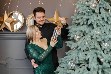 Genç bir kadın ve erkek Noel ağacının yanında duruyor ve sarılıyorlar. Mutlu bir aile Noel 'i kutluyor. Yeşil elbiseli sarışın kadın ve siyah tişörtlü sakallı adam..