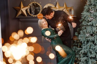 Genç bir kadın ve bir adam yılbaşı ağacının yanında durup maytap tutuyorlar. Mutlu bir aile Noel 'i kutluyor. Yeşil elbiseli sarışın kadın ve siyah tişörtlü sakallı adam..