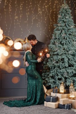 Genç bir kadın ve bir adam yılbaşı ağacının yanında duruyorlar ve ellerinde maytap var. Mutlu bir aile Noel 'i kutluyor. Yeşil elbiseli sarışın kadın ve siyah tişörtlü sakallı adam..