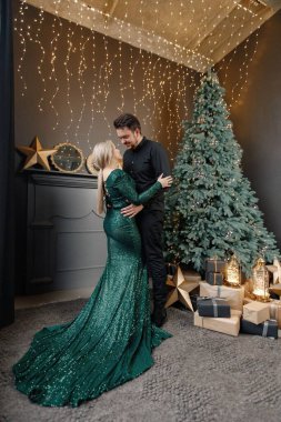 Genç bir kadın ve erkek Noel ağacının yanında duruyor ve sarılıyorlar. Mutlu bir aile Noel 'i kutluyor. Yeşil elbiseli sarışın kadın ve siyah tişörtlü sakallı adam..