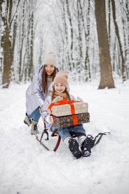 Anne ve çocuğu, kar tatilinde ormanda eğleniyorlar. Mor kalın ceketler ve örgü şapkalar giyen kızlar. Hediye kutusu tutan bir kız..