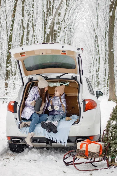 Young brunette woman sitting in open cars trunk with her daughter and holding a thermos with tea. Female models posing for a photo. Family wearing purple warm jackets and knitted hats.