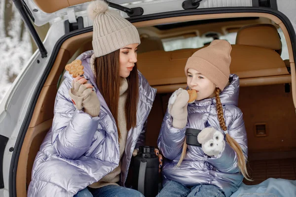 Young brunette woman sitting in open cars trunk with her daughter and eating a cookies. Female models posing for a photo. Family wearing purple warm jackets and knitted hats.