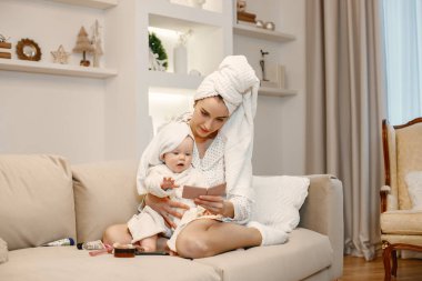 Genç anne ve küçük kız havluya sarılı sabahlıklar giyiyorlar. Kanepede oturan kadın ve kız. Kadın ve kız küçük aynaya bakıyor..