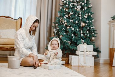 Esmer anne ve küçük kız Noel ağacının yanında oturuyor. Beyaz sabahlık giyen kızlar. Kadın ve kız süs olsun diye top oynuyorlar..