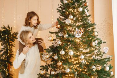 Noel 'i annesiyle birlikte kutlayan küçük bir kızın portresi. Kız ve annesi Noel ağacı süslüyorlar. Bej örgülü giysiler giyen bir aile..