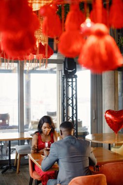 Restoranda eğlenen bir çift. Siyah adam ve kadın kırmızı şarap içip kek yiyorlar. Kırmızı şık elbiseli kadın ve mavi kostümlü adam..