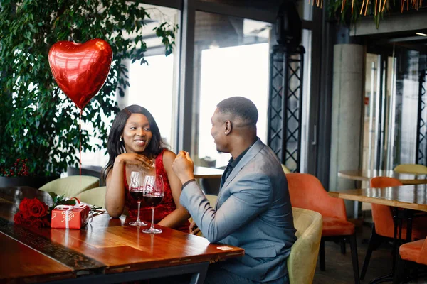 Couple enjoying day out at restaurant. Black man and woman drinking wine. Woman wearing red elegant dress and man blue costume.